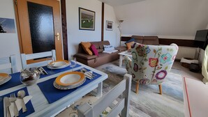 A dining area with a table set for two, a brown sofa, a floral armchair, and a television in the background. - Apartment 'Inselstern' with Balcony and Wi-Fi (Pellworm)