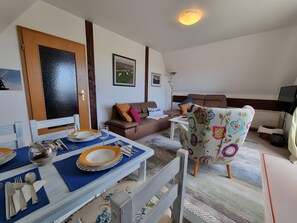 Ein Essbereich mit Tisch für zwei Personen, einer braunen Couch, einem floralen Sessel und einem Fernseher im Hintergrund.