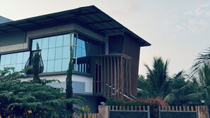 Una casa moderna de dos pisos con ventanas amplias, entrada protegida por un portón y abundante vegetación alrededor.