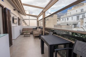 A modern balcony with a glass enclosure, beige sofas, a dining table, and a view of the cityscape. - Athens Luxury Penthouse (Athens)