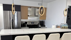 A modern kitchen with a stainless steel refrigerator, a built-in oven, and a coffee machine. There are three bar stools with a light-colored cushioned seat.