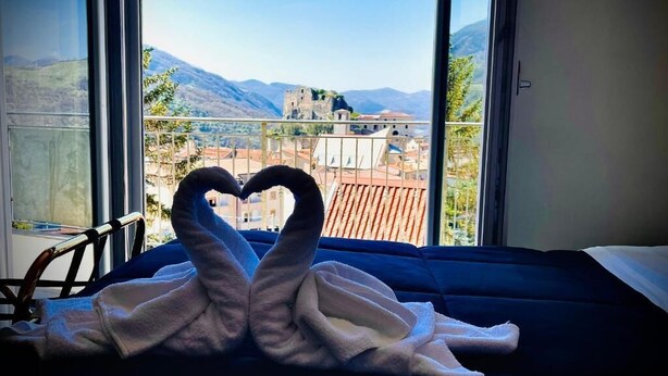A bed with a heart-shaped towel arrangement, a window with a view of a historic building and mountains. - Beb Villa Real (Balvano)