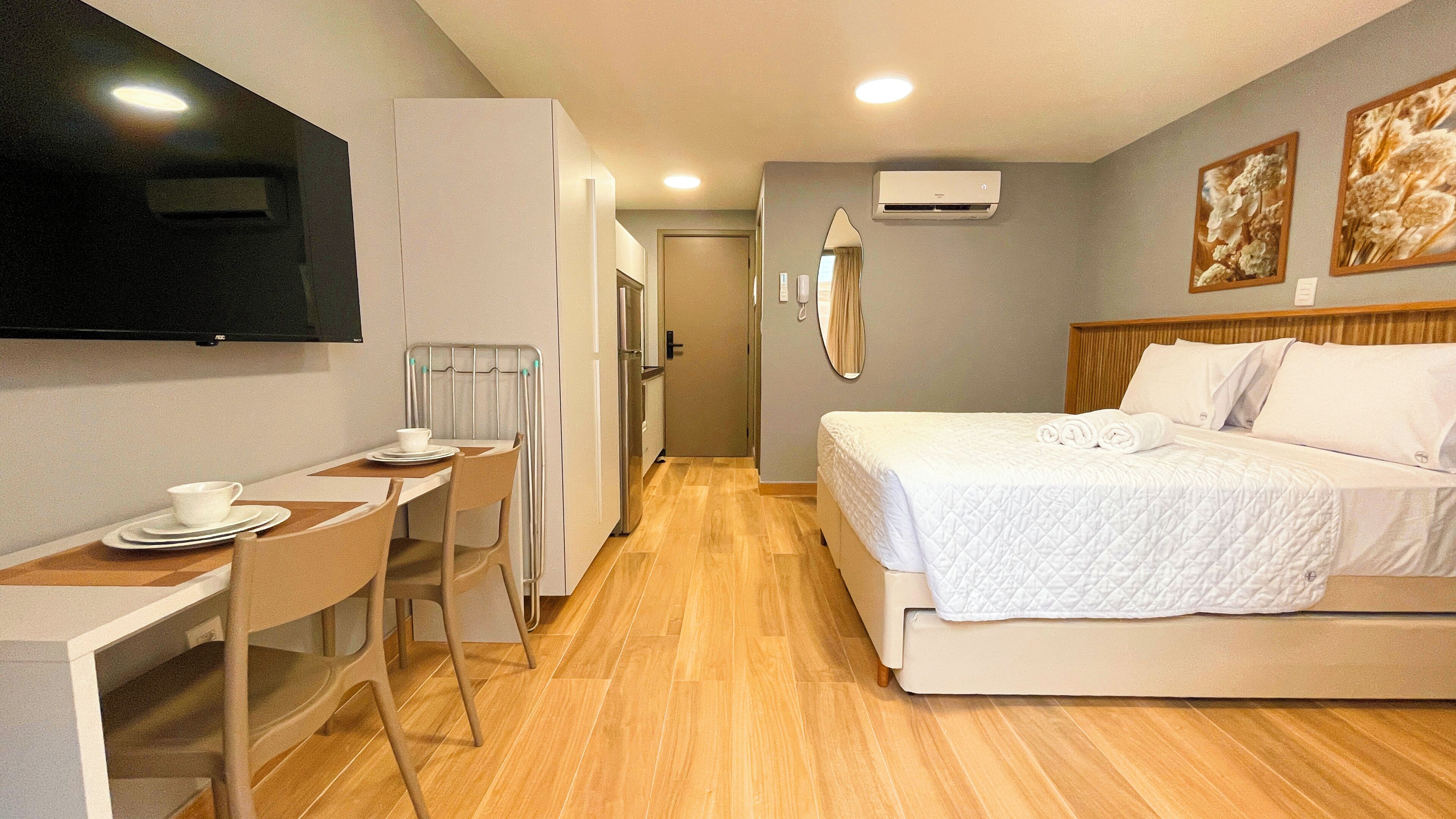 A hotel room with a bed, a TV mounted on the wall, a desk with a chair, and a small table with a cup and plates.