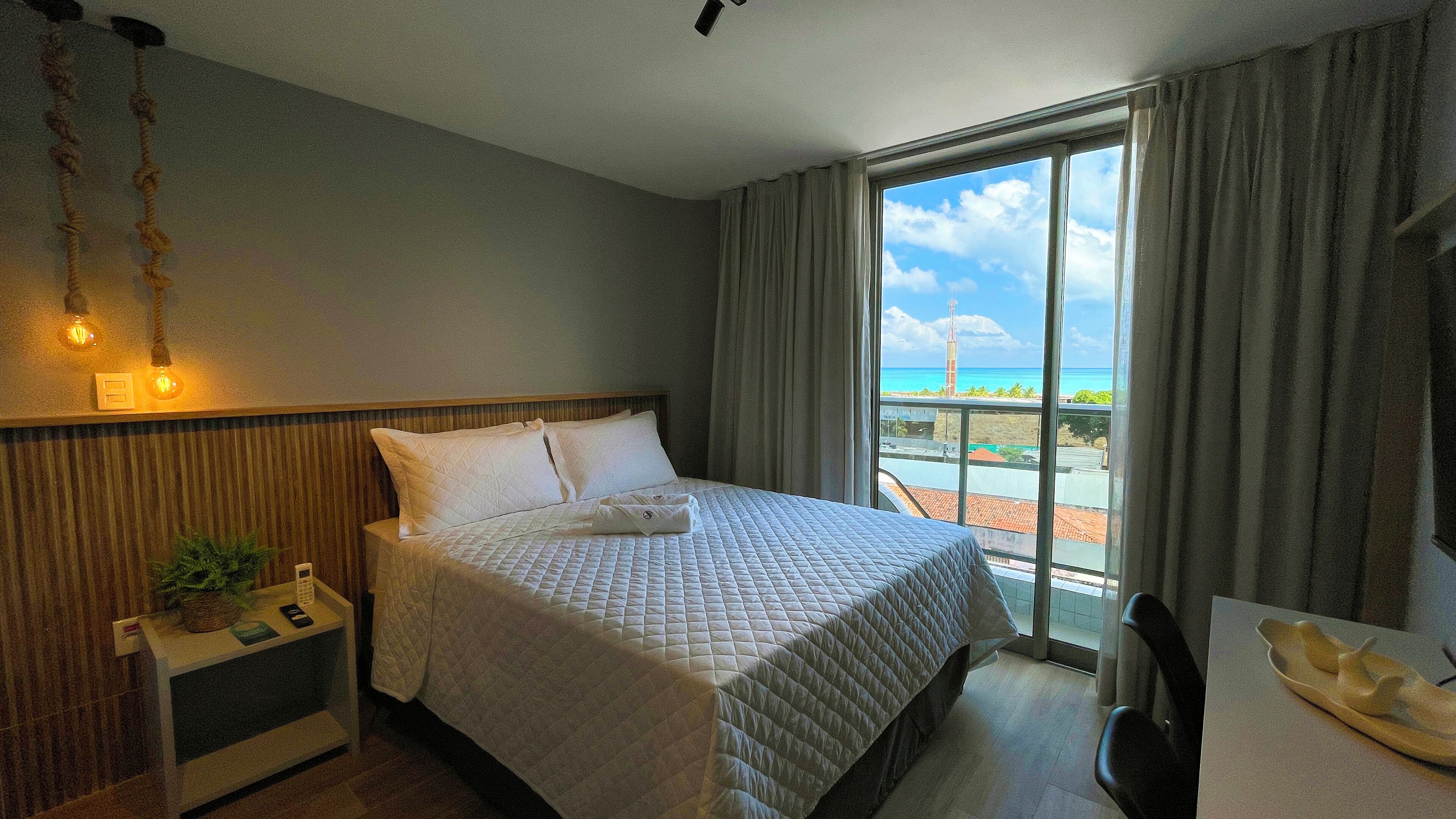 A hotel room with a bed, a nightstand, a desk with a chair, a window with a view of the beach, and a ceiling light fixture.