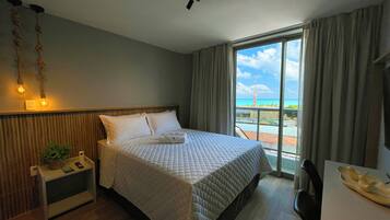 A hotel room with a bed, a nightstand, a desk with a chair, a window with a view of the beach, and a ceiling light fixture.