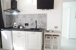 Eine kompakte Küche mit weißen Schränken, einer schwarzen Dunstabzugshaube, einer strukturierten Spritzschutzwand und einem kleinen weißen Kühlschrank.