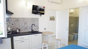 Een kleine keuken met witte kasten, een zwart aanrecht en een betegelde spatwand. Er staan twee barkrukken en een klein tafeltje. Een poster hangt aan de muur, en een deur leidt naar de badkamer.