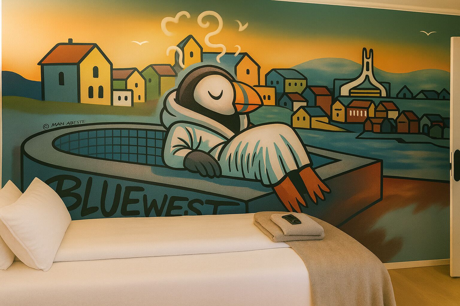 Ein Zimmer mit einem Bett, einem Wandbild eines schlafenden Pinguins in einer Badewanne und dem Schriftzug "BLUEWEST" auf der Wanne.