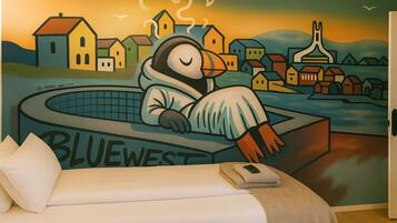 Ein Zimmer mit einem Bett, einem Wandbild eines schlafenden Pinguins in einer Badewanne und dem Schriftzug "BLUEWEST" auf der Wanne.