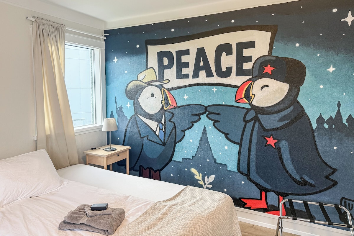 A hotel room with a bed, a nightstand, a lamp, and a wall mural featuring two birds with a 'PEACE' banner.