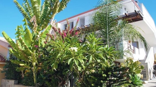 A white building with a red roof and a balcony. There are various plants and trees in front of the building. - Sirocco - House of Jana (Quartu Sant'Elena)