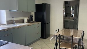 A modern kitchen with grey cabinets, a black refrigerator, a dining table with chairs, and a tiled floor. - Beautiful studio (Volx)