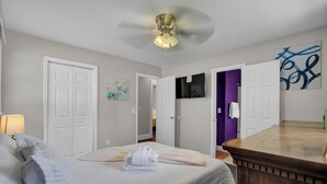 Ein Schlafzimmer mit Bett, Nachttisch, Deckenventilator, Badezimmer mit Toilette und Waschbecken sowie ein Flur mit einer Tür zu einem weiteren Raum.