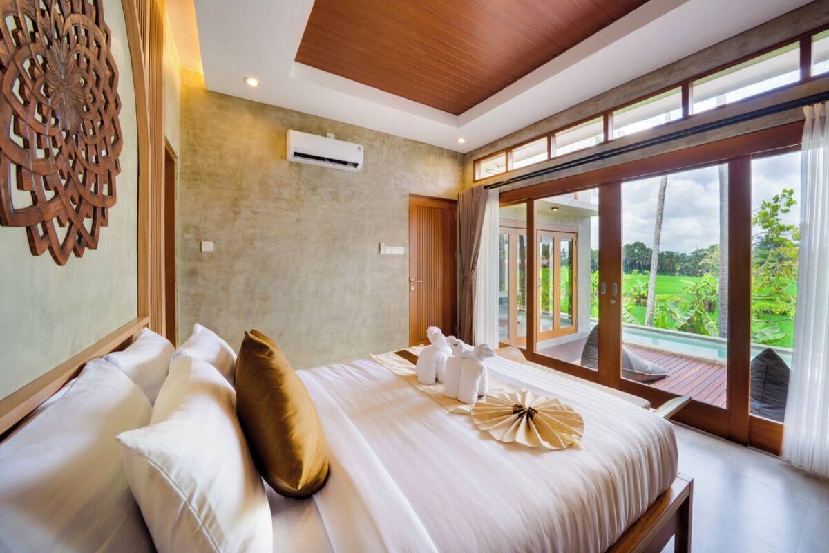 A large bed with white linens and a decorative pillow, a wooden ceiling, and a view of a lush garden through large windows.