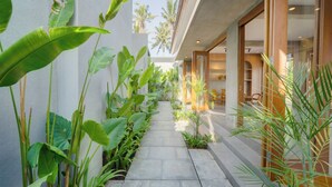 Un vialetto stretto fiancheggiato da grandi piante tropicali che conduce a un edificio con ampie vetrate e un design moderno.