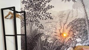 Une pièce ornée d’une fresque représentant un paysage naturel, avec une étagère noire, un petit tabouret en bois et un lit recouvert d’une couverture rose.