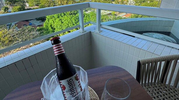 Varanda com mesa posta para dois, uma garrafa de champanhe e vista para o oceano e vegetação.