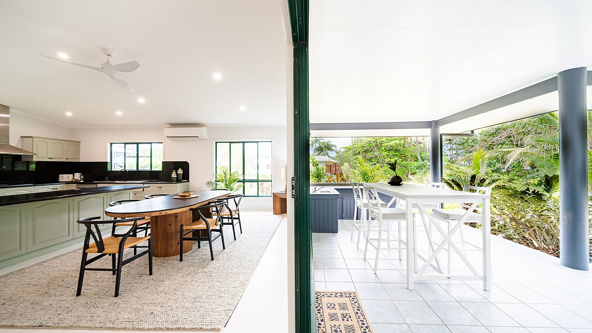 A modern kitchen with a central island, dining area with white chairs, and a view of lush greenery outside.