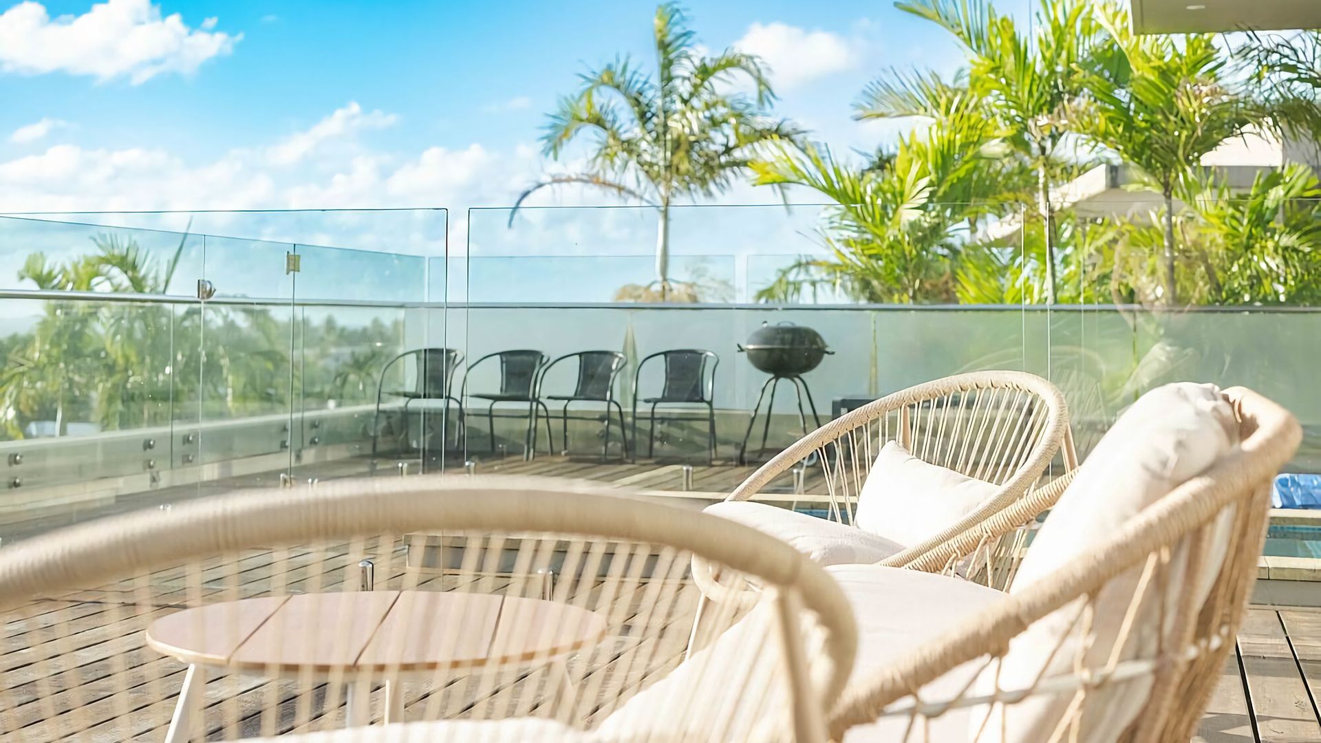A glass-enclosed outdoor seating area with wicker chairs and a round table, overlooking a pool and palm trees.