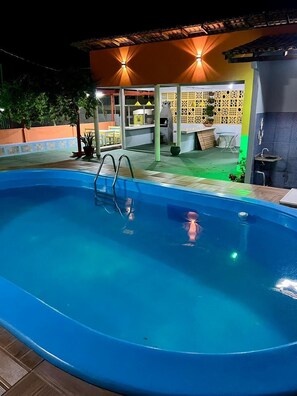 Uma piscina com uma escada de metal, rodeada por um terraço com um espaço de estar coberto e uma cerca decorativa.