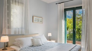 Ein Schlafzimmer mit Bett, Nachttischen, einer Lampe, einem gerahmten Bild an der Wand und Blick auf Grün durch das Fenster.