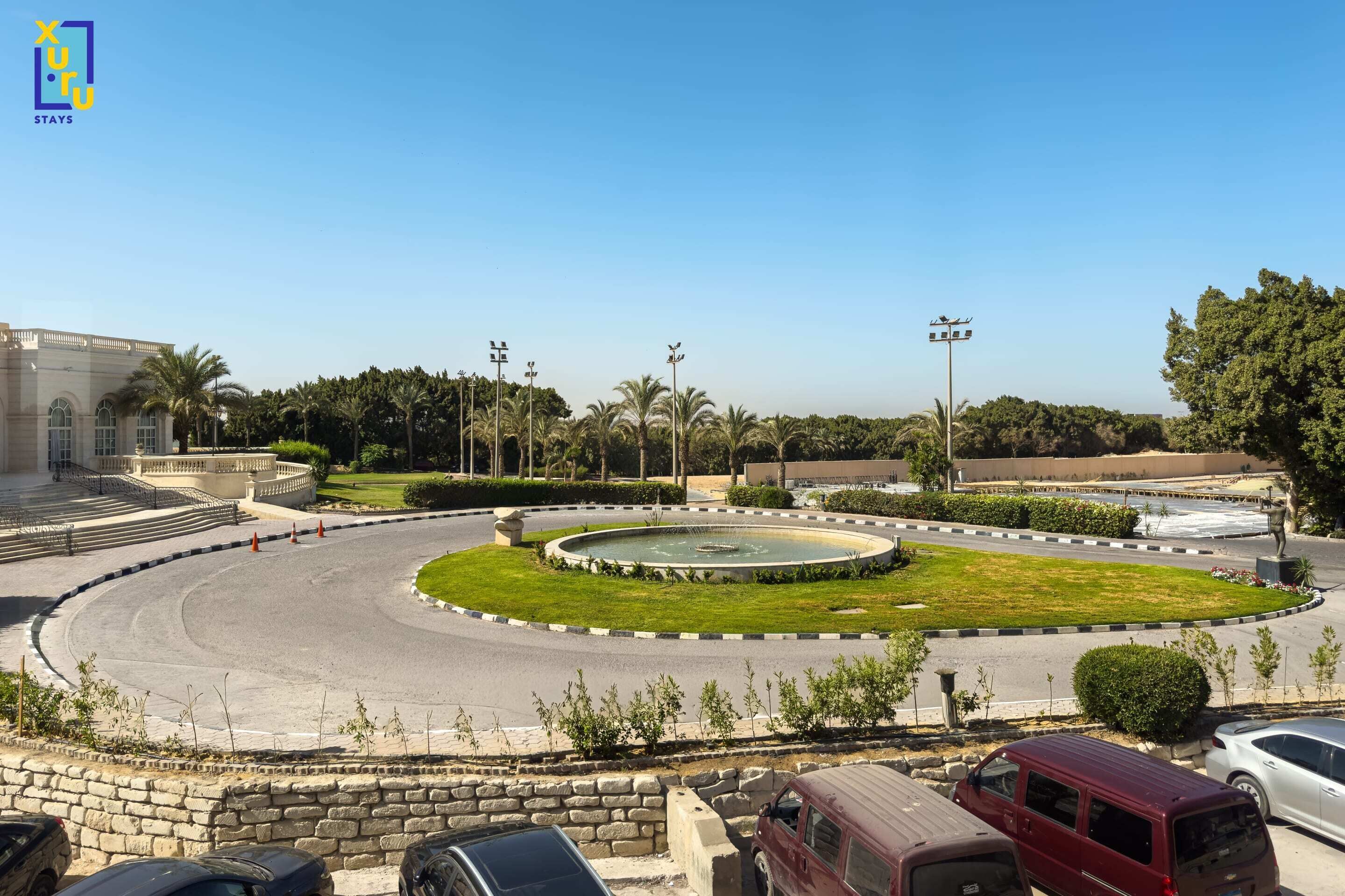 A roundabout with a central fountain, palm trees, and a stone wall.