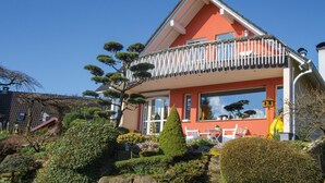 Een huis met een balkon en een tuin vol planten, onder een heldere blauwe lucht.
