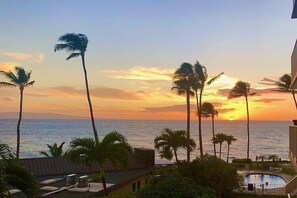 A sunset view from a balcony overlooking a swimming pool, palm trees, and the ocean. - Kamaole Nalu 201 - Updated bright 2 bedroom ocean front (Kihei)