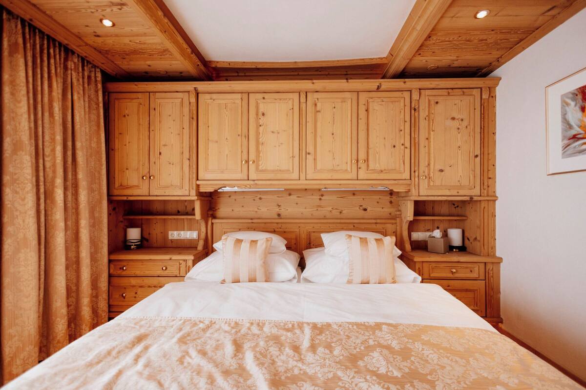 Un dormitorio con cabecera de madera, cama con ropa de cama blanca, mesitas de noche de madera y un ventanal con cortinas.