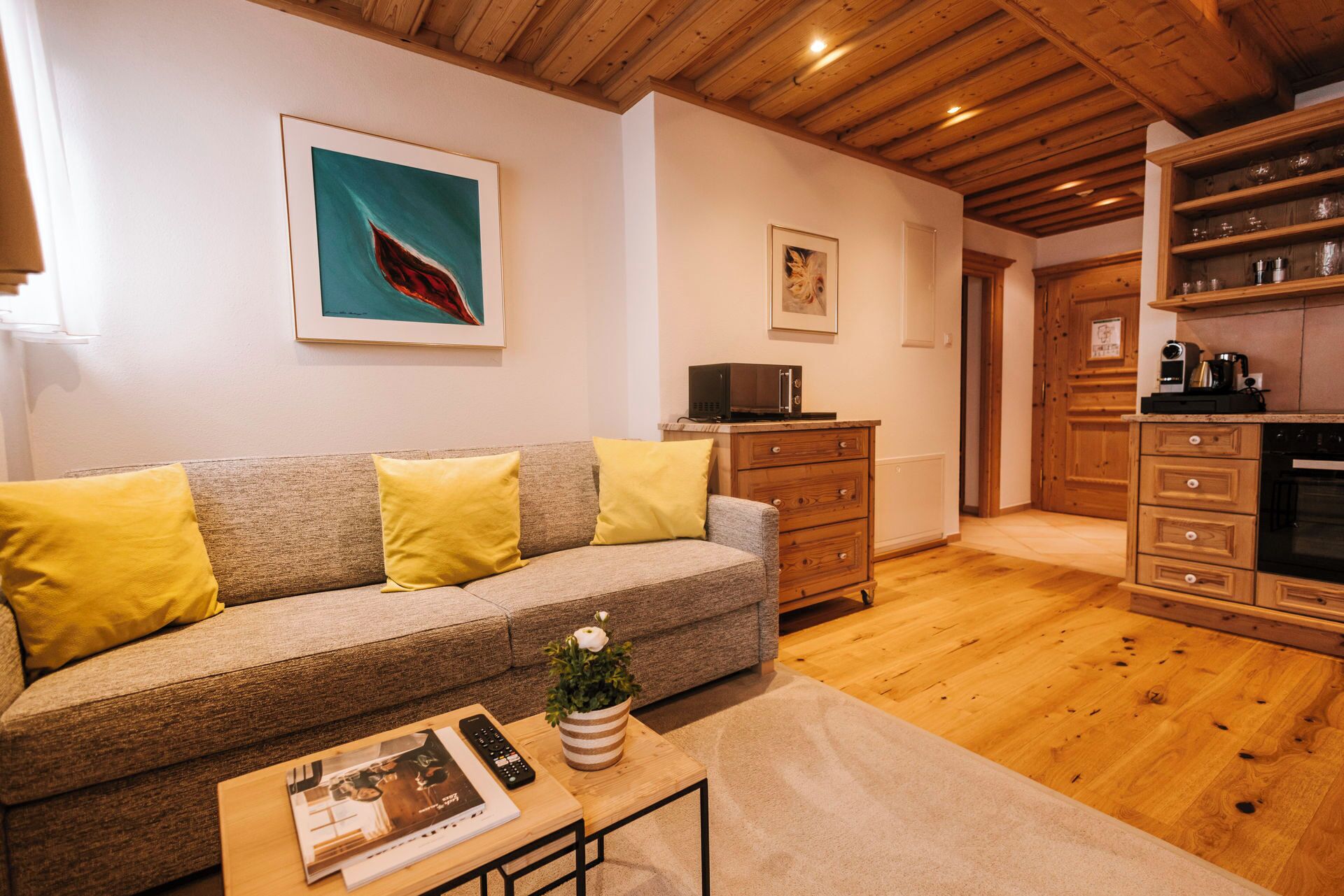 A cozy living room with a sofa, a coffee table with a book and remote, a plant, and a kitchen area with wooden cabinets and appliances.