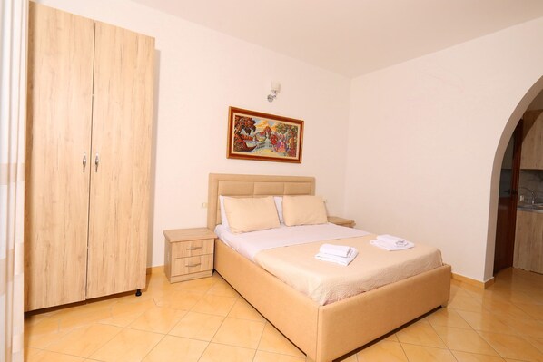 A bedroom with a bed, a wardrobe, a nightstand, a painting on the wall, and a bathroom visible through an archway. - Hill Suites (Vlore)