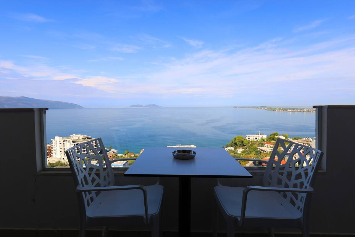 Varanda com mesa e duas cadeiras, oferecendo vista da costa, com edifícios e céu claro.