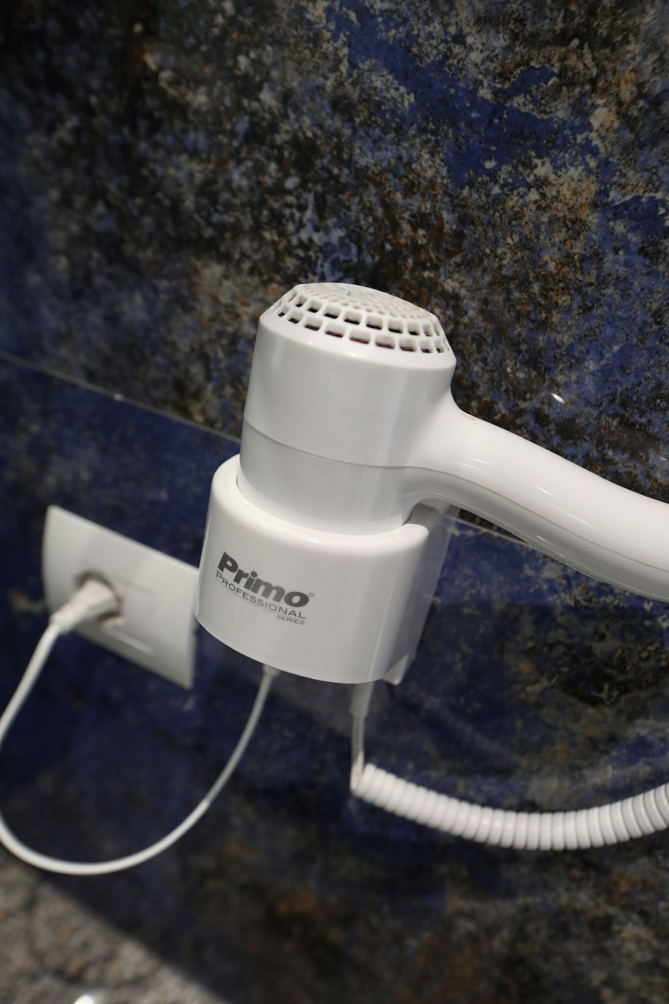 A white hair dryer with the brand "Primo Professional Series" attached to a wall socket.