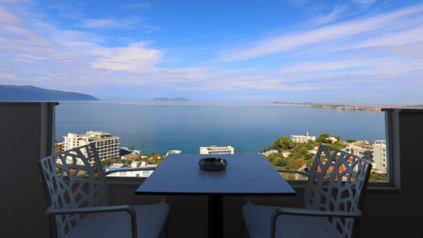 A balcony with a table and chairs overlooking a body of water and cityscape. - Hill Suites (Vlore)
