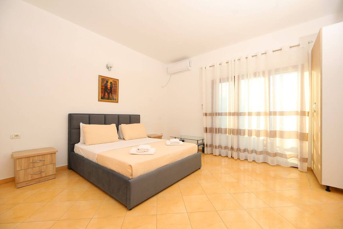 Quarto com cama, mesas de cabeceira, ar condicionado fixado na parede e janela com cortinas.