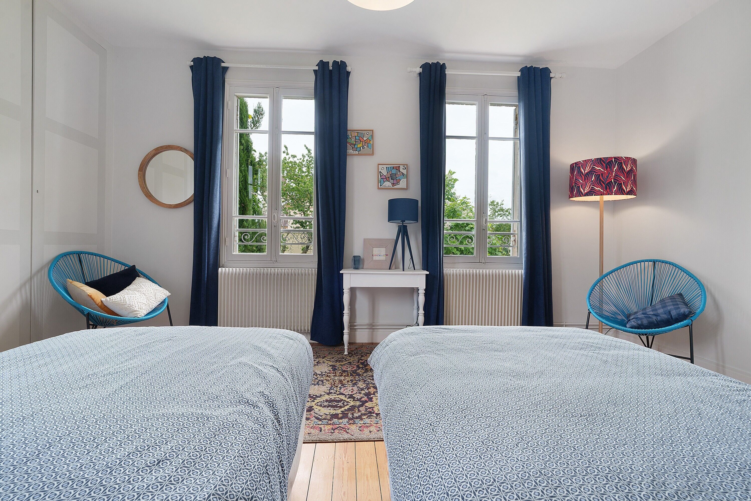 A room with two beds, a small table, a chair, a lamp, and a view of greenery through the windows.