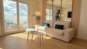 Un salon moderne avec un canapé beige, une petite table ronde et une grande porte-fenêtre coulissante donnant sur une terrasse extérieure où se trouve une chaise bleue.