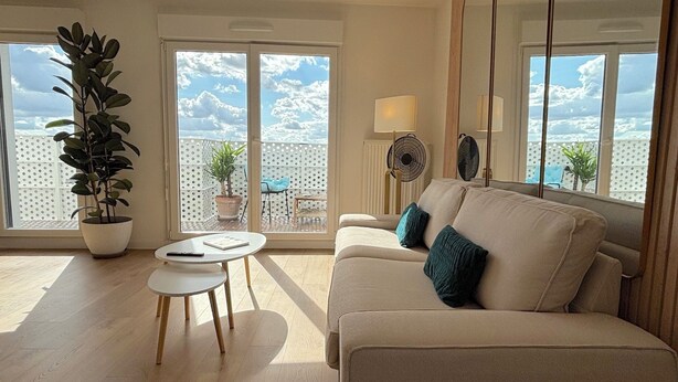 A modern living room with a beige sofa, two teal cushions, a small white table, and a potted plant. - Le Penthouse Pesaro C - Concert Celine DION (Nanterre)