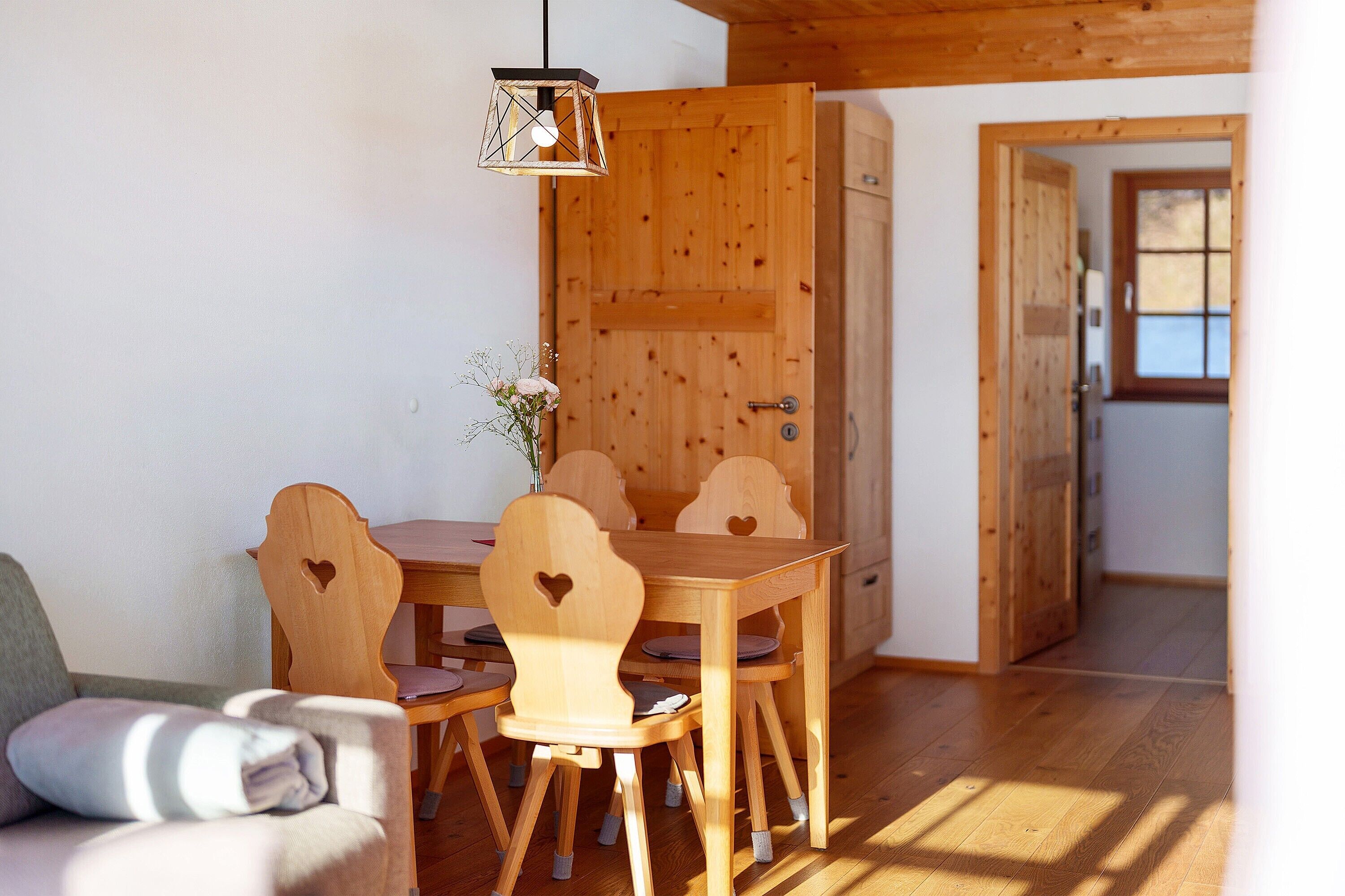 Ein Esszimmer mit Holztisch, Stühlen, einer Couch und einer Pendelleuchte. Die Wände und der Boden sind ebenfalls aus Holz.