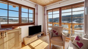 A room with large windows, a wooden ceiling, a TV on a wooden cabinet, and a chair with heart-shaped pillows. - Apartment 'Hirtebube' with Shared Garden, Balcony, and Wi-Fi (Lenzkirch)