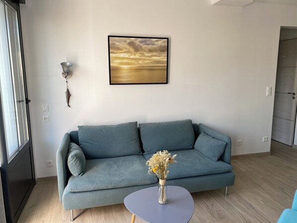 Een woonkamer met een grijze bank, een vaas met bloemen, een lijst met een zonsondergang en een deur rechts.