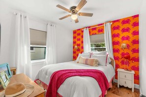 Une chambre à coucher comprenant un lit, un ventilateur de plafond, une fenêtre avec des rideaux, une table de chevet et un mur orné d’un motif.