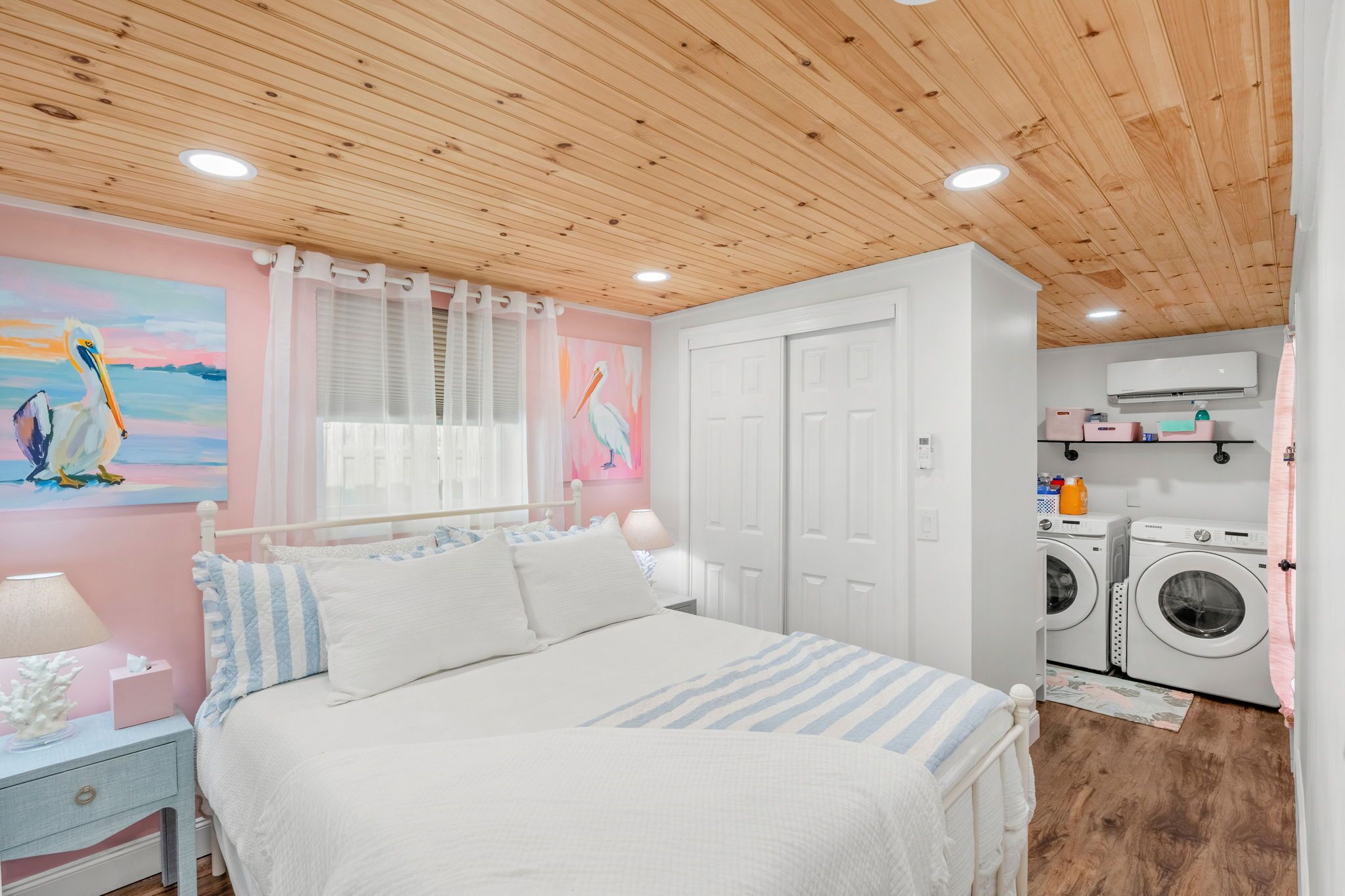A bedroom with a wooden ceiling, a bed with white bedding, a nightstand with a lamp, a wall with a painting of birds, and a laundry area with washing machines.