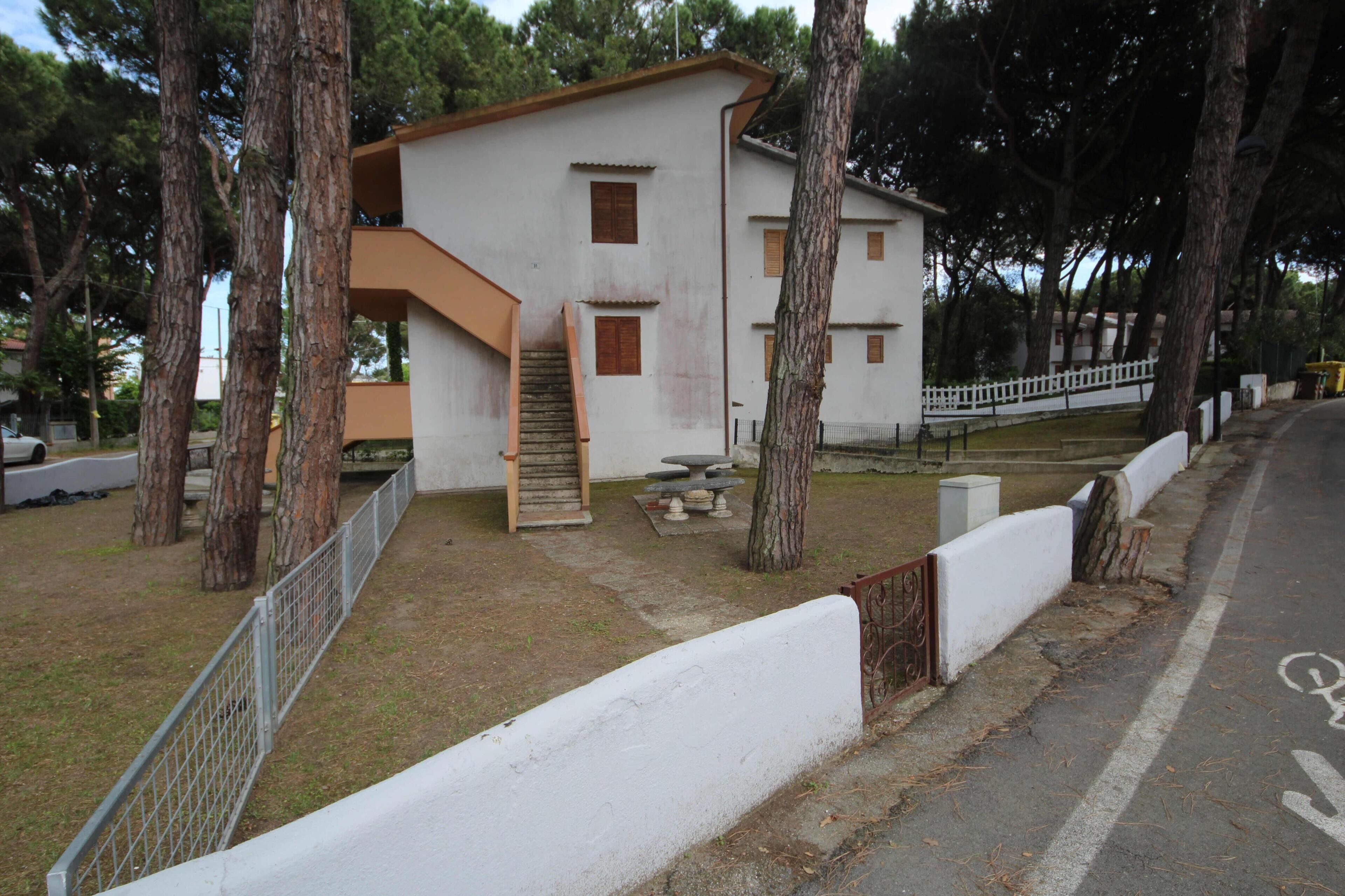 A two-story building with a staircase leading to the upper floor, surrounded by trees and a fenced area.