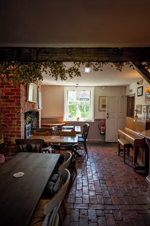 A brick-floored dining area with a fireplace, wooden tables and chairs, a window with a view of greenery, and a door with a fire extinguisher. - The Sportsman Inn (Amberley)