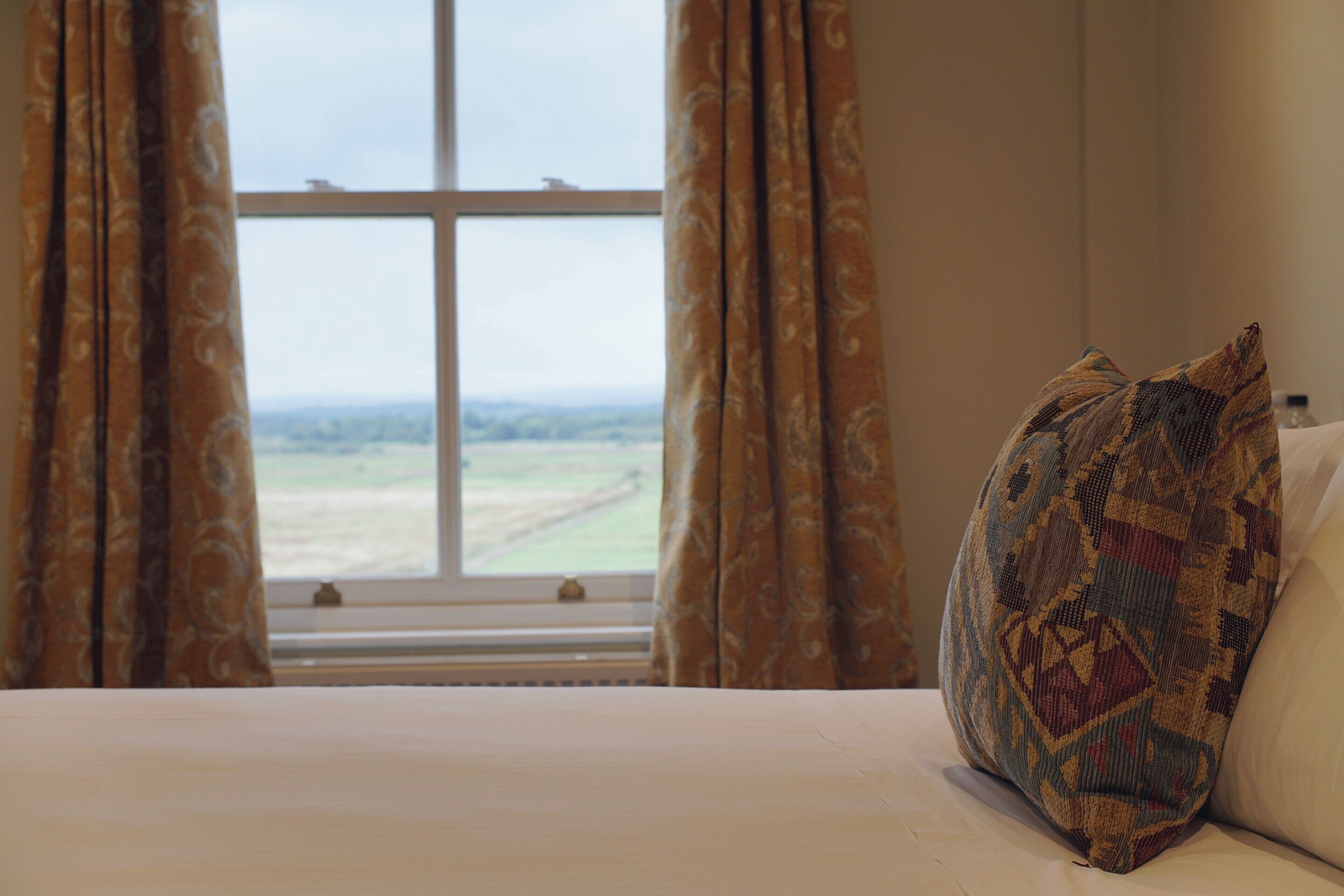 Una cama con un cojín estampado y vista a un paisaje a través de una ventana con cortinas.