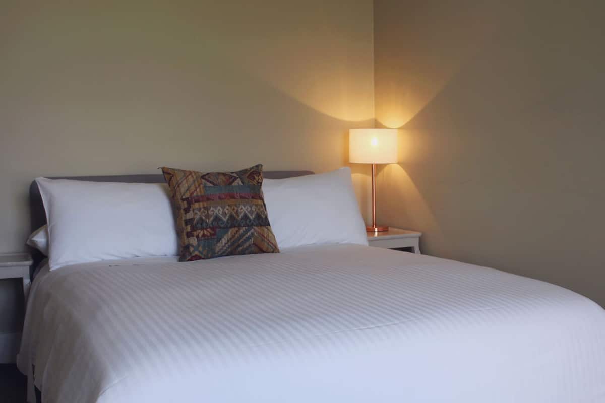 A neatly made bed with white linens and a patterned pillow, a bedside lamp providing soft light, and a plain wall in the background.