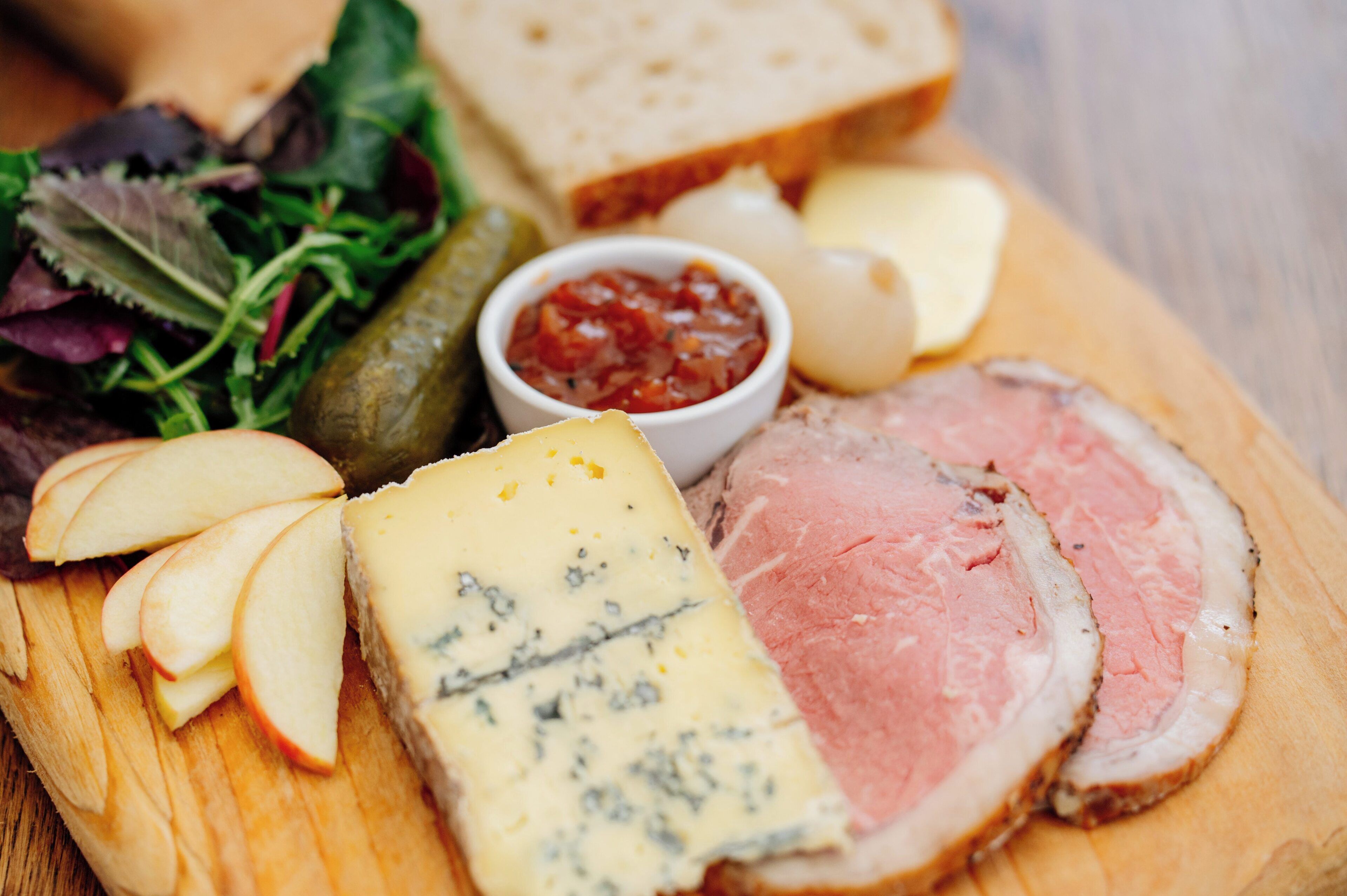 Una tabla de madera con jamón en rodajas, queso, manzanas, pepinillos, pan y una ensalada.