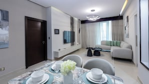 A modern living room with a sofa, a dining table set for four, and a TV mounted on the wall. - Stylish 2BR Business Bay Pool Access (Dubai)
