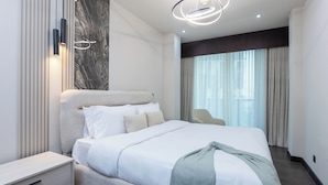 A modern hotel room with a large bed, a bedside table, a wall-mounted light fixture, and a view of the outdoors through a window. - Stylish 2BR Business Bay Pool Access (Dubai)